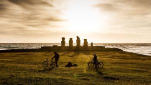 A scenic view of Easter Island during sunset with two people riding bicycles across a grassy field against the Moai statues in the background silhouetted against the golden sky