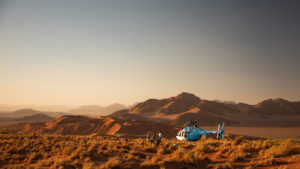 A small helicopter parked in the vast orange-hued Sossusvlei desert landscape with rolling dunes and a group of people nearby